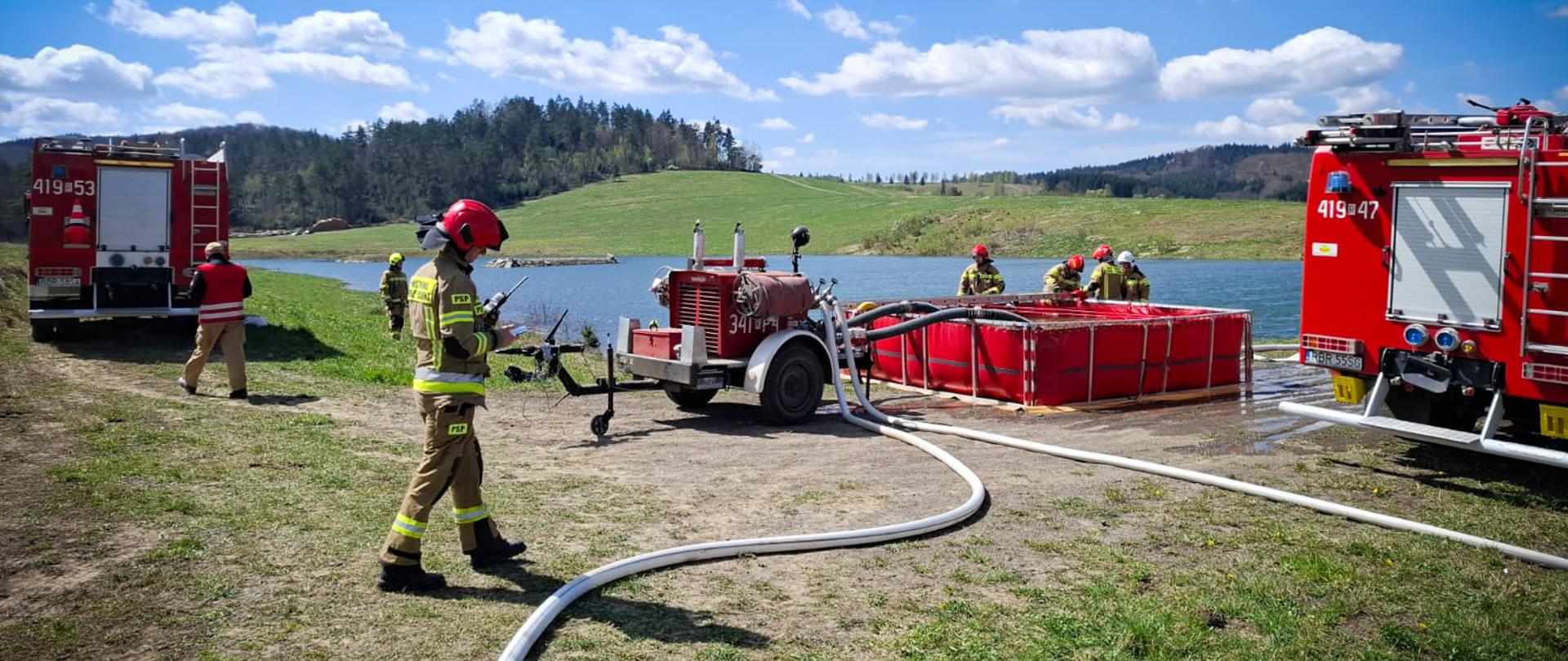 Strażacy przygotowują punkt poboru wody nad brzegiem zbiornika wodnego. Na pierwszym planie widoczny strażak w pełnym umundurowaniu bojowym (nomex) i czerwonym hełmie, idący wzdłuż rozwiniętej białej linii wężowej. W tle znajduje się czerwony, przenośny zbiornik na wodę oraz motopompa na przyczepie. Po bokach stoją dwa ciężkie samochody ratowniczo-gaśnicze.