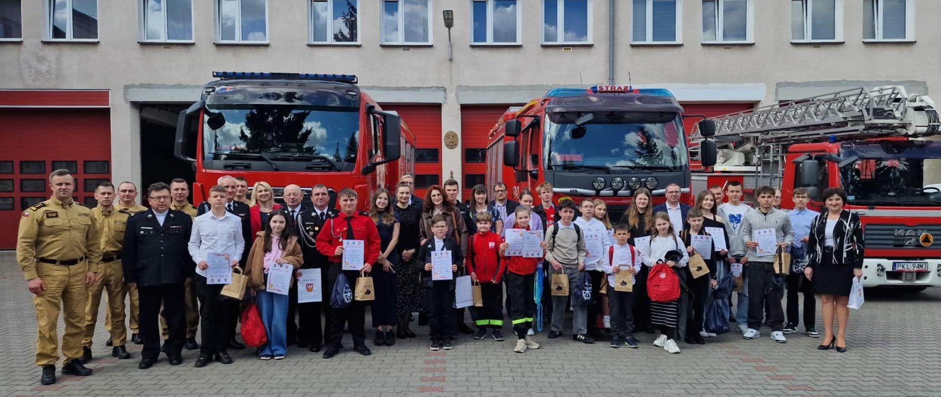 Zdjęcie przedstawia wszystkich uczestników Powiatowych Eliminacji Turnieju Wiedzy Młodzież Zapobiega Pożarom wraz z opiekunami i strażakami na tle wozów strażackich