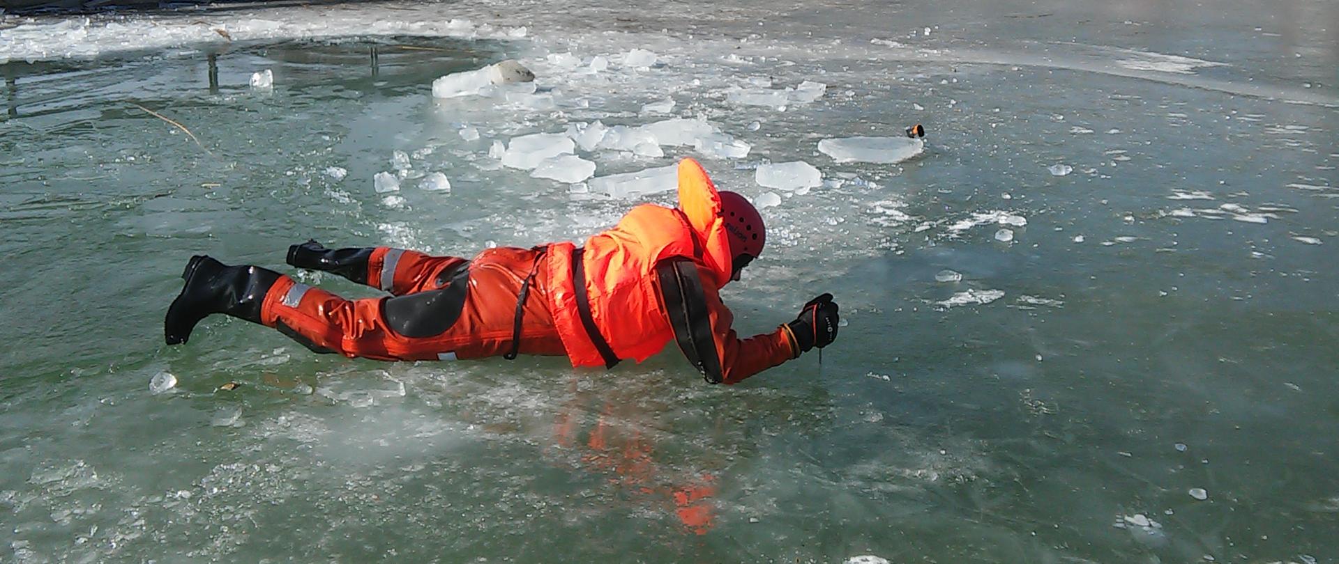 Człowiek próbuje wydostać się z przerębli lodowej. Ubrany jest w skafander wodoszczelny oraz kask i kamizelkę ratunkową. Na drugim planie brzeg akwenu z infrastrukturą.