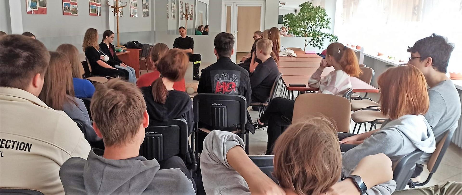 Na zdjęciu przedstawiono warsztaty z uczniami w zespole szkół w ramach profilaktyki bezpieczeństwa. Grupa młodzieży licealnej słucha prelekcji prowadzonej przez ratownika.