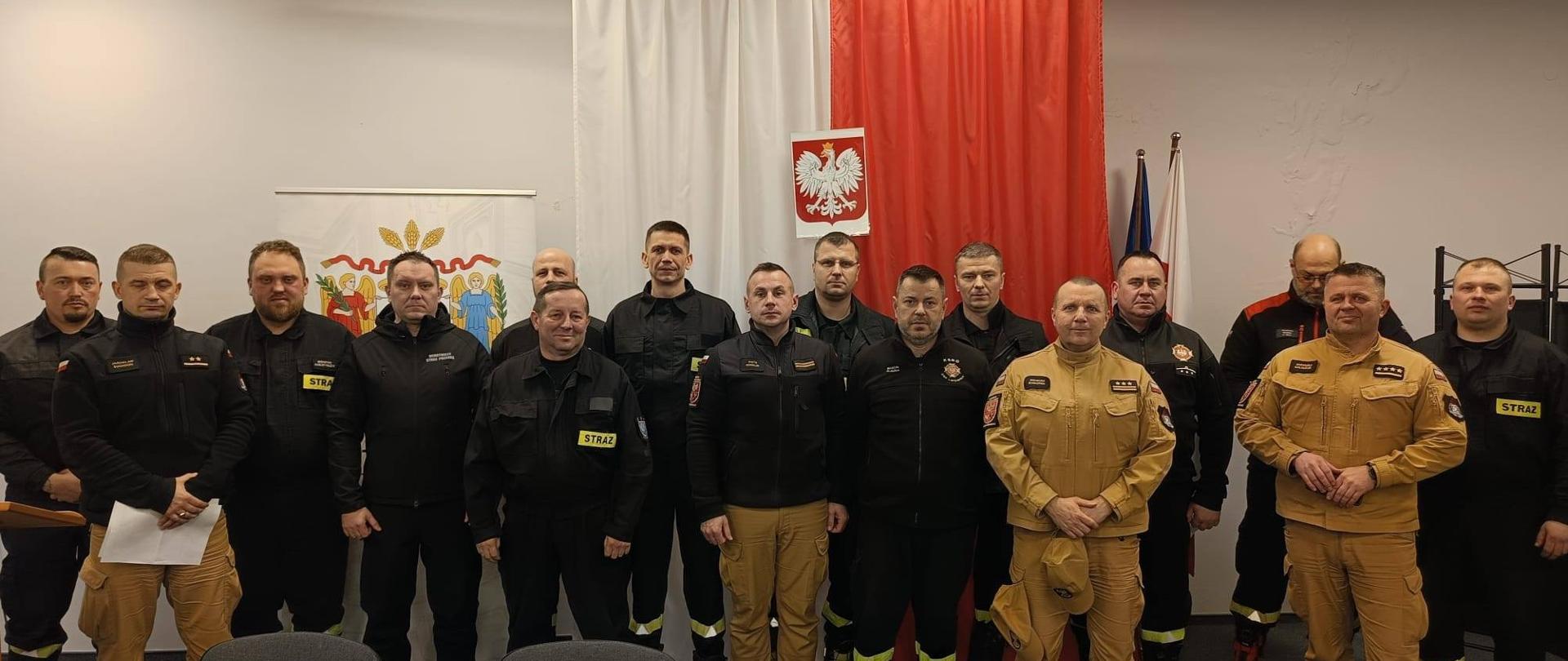 Grupa strażaków w jasnych i czarnych mundurach na sali szkoleniowej. W tle w pionie flaga Polski.