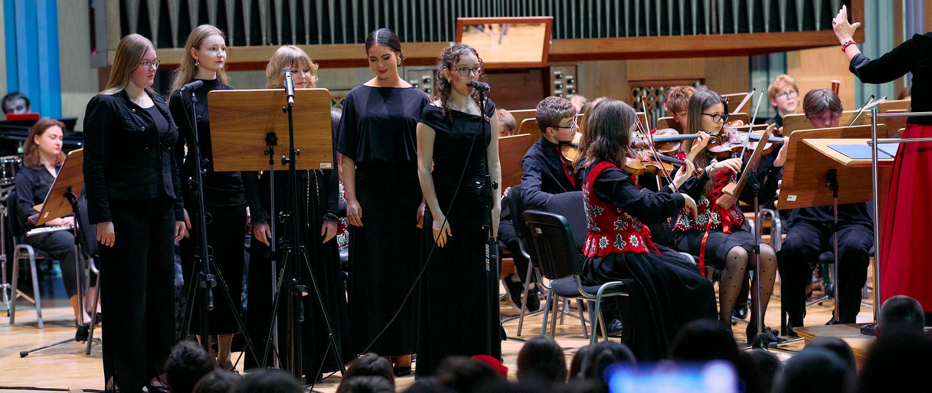 Na scenie sali koncertowej trwa występ muzyczny. Na pierwszym planie, nieco z lewej strony, stoi pięć kobiet w eleganckich, ciemnych strojach — głównie długie czarne sukienki lub czarne komplety. Ustawione są w rzędzie przed statywami z nutami i mikrofonem. Wyglądają na wokalistki, gotowe do śpiewu lub właśnie śpiewające.
Za nimi widać wielką fasadę organów piszczałkowych, z licznymi, równomiernie ułożonymi, błyszczącymi, pionowymi rurami. Po prawej stronie sceny, nieco z tyłu, siedzi orkiestra smyczkowa. W pierwszym rzędzie dwa skrzypaczki zwrócone twarzami w prawo, ubrani w czarne stroje, z czerwonymi kamizelkami ozdobionymi wzorami. Widać ich skupienie i aktywne granie, smyczki w ruchu. Dalej widać więcej muzyków, również grających na smyczkach, oraz stojącą po prawej krawędzi zdjęcia osobę — najpewniej dyrygenta — unoszącą rękę, jakby prowadziła orkiestrę. Dyrygent ubrany jest w czerwony dół stroju, choć tylko fragment jest widoczny.
Na dole zdjęcia, w ciemniejszym obszarze, częściowo widoczna jest publiczność. W pierwszym rzędzie ktoś trzyma telefon lub mały ekran, na którym widać rozmyty, jasny punkt światła, jakby nagrywał występ. Głowy widzów tworzą ciemny, niewyraźny kształt przeciw kontrastowi jasnej sceny.
