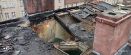 Zdjęcie przedstawia poszycie dachu budynku mieszkalnego, który objęty był pożarem. W dachu przy kominie znajduje się otwór pokazujący wnętrze klatki schodowej. W tle znajdują się budynki usytuowane na ulicy Śląskiej.