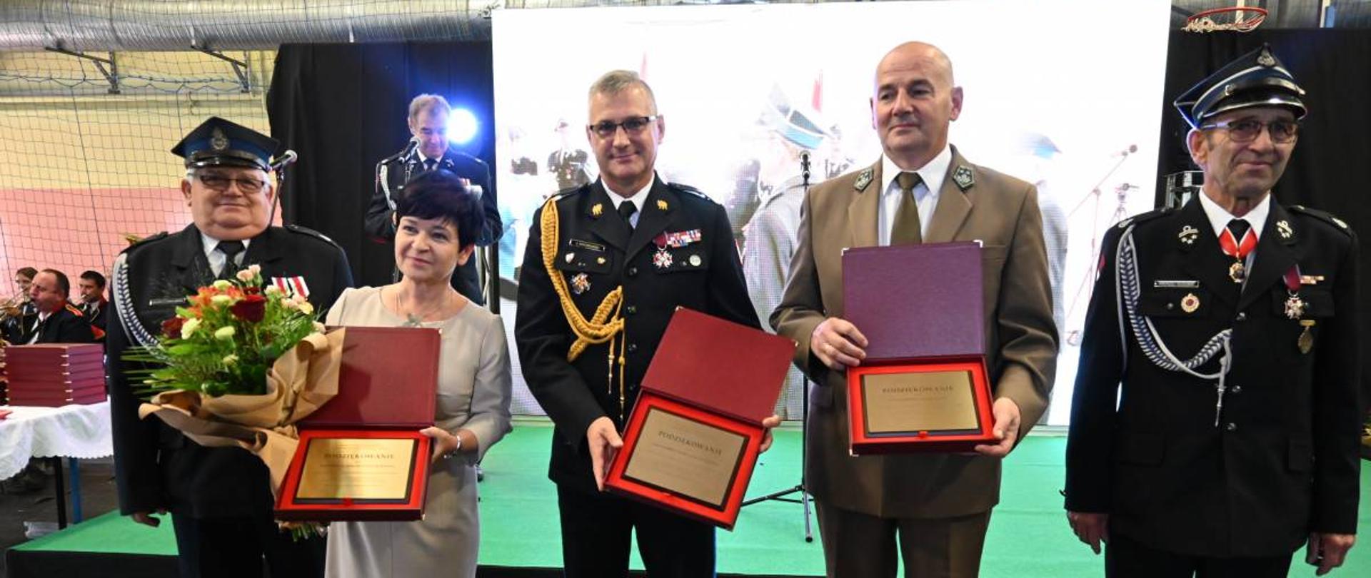 w szeregi stoą wyróżnione osoby, obdarowane okolicznościowym upominkiem. Od lewej: druh OSP. Pani Poseł, komendant wojewódzki, przedstawiciel Lasów Państwowych i kolejny druh OSP. W tle ekran.