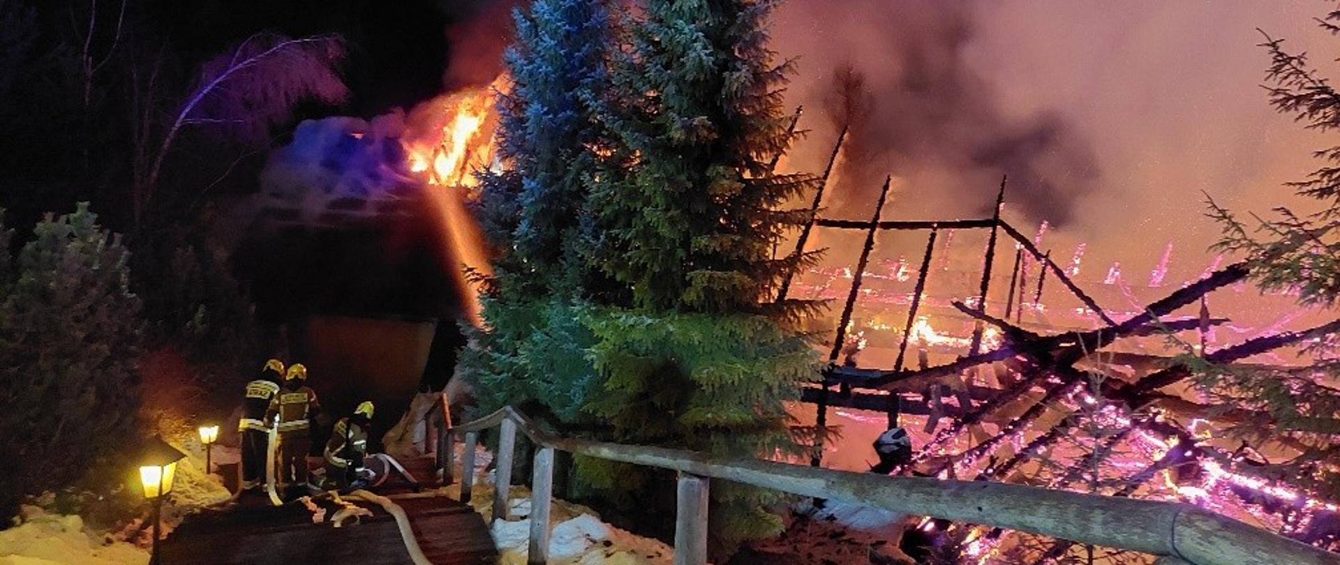 Noc. Po prawej stronie płonący drewniany budynek. Centralnie schody. W dole 3 strazaków. Widoczne węże pożarnicze oraz strumien wody. Na środku zdjęcia choinki.