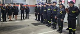 W prawej części zdjęcia Strażacy w mundurach wyjściowych i służbowych stoją w dwuszeregu na tle bram garażowych, w centralnej i lewej jego części Komendant odczytuje treść z tabliczki okolicznościowej, obok niego zastępca Komendanta i inni pracownicy Komendy.