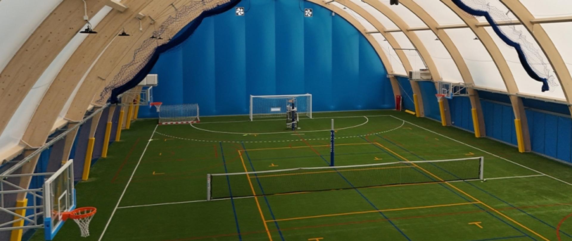 Na zdjęciu widać wnętrze hali sportowej. Na trawiastej nawierzchni namalowane są linie, które wyznaczają boisko. No końcu boiska stoi bramka.