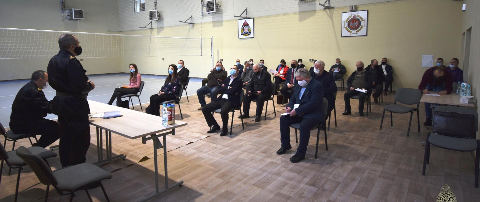 Grupa ludzi siedzących na krzesłach, z lewej dwóch strażaków (tyłem do fotografującego), jeden stoi, drugi siedzi.