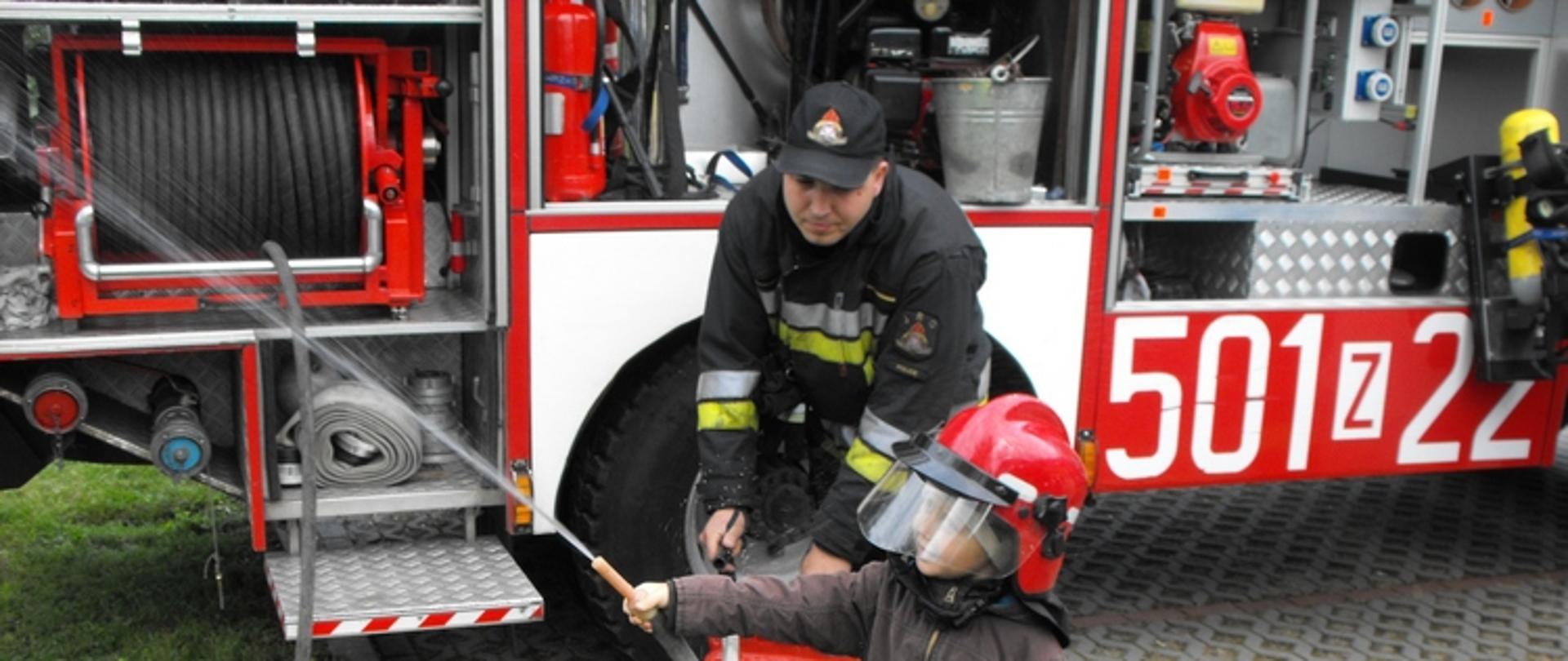 Zdjęcie przedstawia małe dziecko trzymające prądowniczkę hydrnetki, stojący strażak pomaga lać wodę 