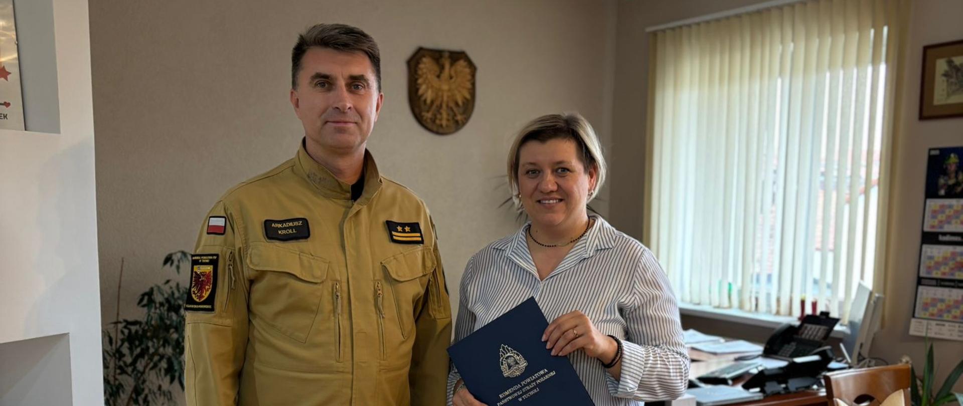 Dwie osoby stoją obok siebie. Z lewej strony stoi Komendant Powiatowy PSP w Tucholi. Komendant ma na sobie umundurowanie koloru piaskowego. Z prawej strony stoi kobieta. Kobieta trzyma w ręku granatową teczkę z logiem Państwowej Straży Pożarnej. Kobieta ubrana jest w białą koszulę w niebieskie paski oraz niebieskie spodnie. W tle widać ścianę. Na ścianie wisi godło Polski. Z tyłu za kobietą widać drewniane biurko z urządzeniami biurowymi. Po prawej stronie na ścianie widać okno oraz stół z krzesłami. Z lewej stronie na ścianie widać obrazy.