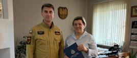 Dwie osoby stoją obok siebie. Z lewej strony stoi Komendant Powiatowy PSP w Tucholi. Komendant ma na sobie umundurowanie koloru piaskowego. Z prawej strony stoi kobieta. Kobieta trzyma w ręku granatową teczkę z logiem Państwowej Straży Pożarnej. Kobieta ubrana jest w białą koszulę w niebieskie paski oraz niebieskie spodnie. W tle widać ścianę. Na ścianie wisi godło Polski. Z tyłu za kobietą widać drewniane biurko z urządzeniami biurowymi. Po prawej stronie na ścianie widać okno oraz stół z krzesłami. Z lewej stronie na ścianie widać obrazy.