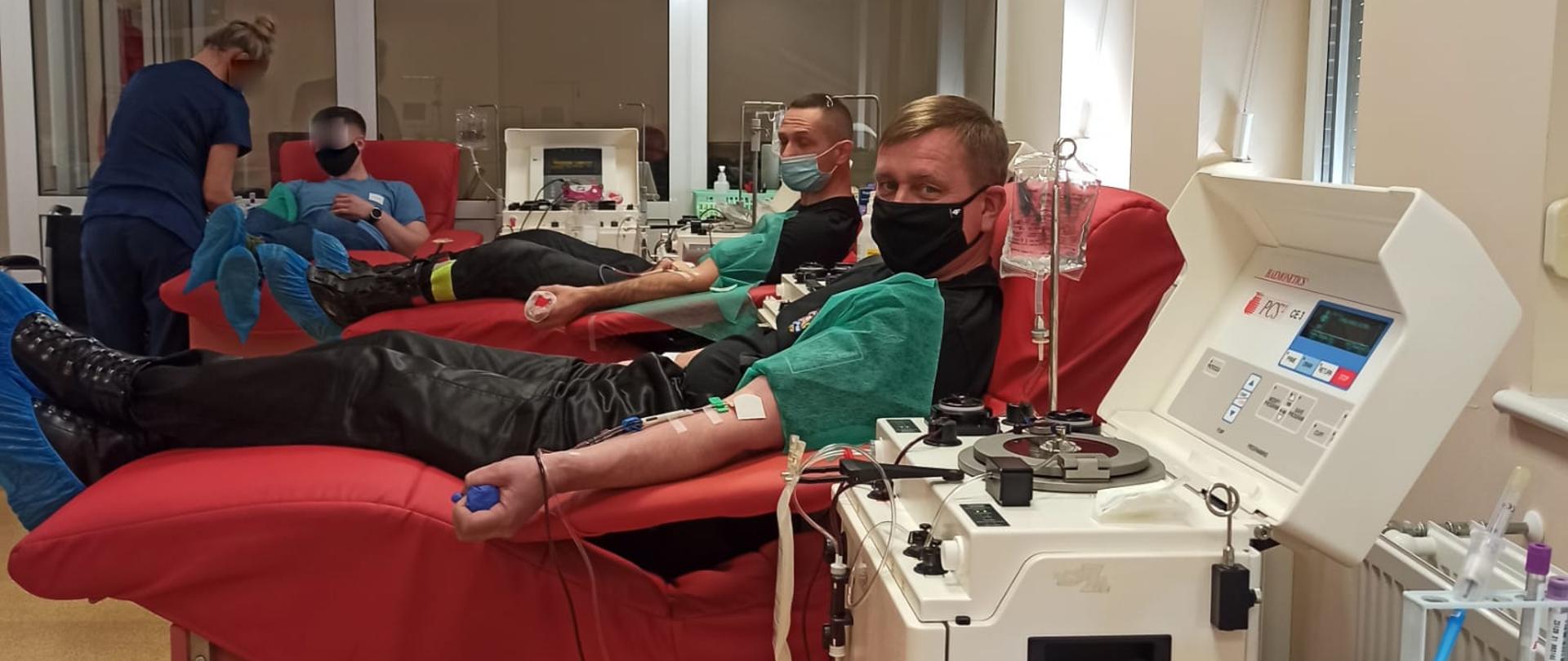 Na zdjęciu widać dwóch strażaków w czarnych mundurach, którzy siedzą na fotelu i oddają osocze. W tle pielęgniarka przygotowuje kolejnego dawcę do oddania osocza.