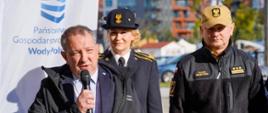 Mężczyzna trzymając mikrofon przemawia na tle baneru Państwowego Gospodarstwa Wodnego Wody Polskie. Za nim stoją funkcjonariusze Państwowej Straży Pożarnej. 