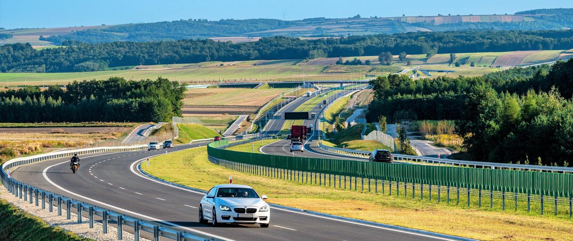 Droga ekspresowa S7 na odcinku Moczydło - Miechów. Dwujezdniowa droga ekspresowa wijąca się między polami i lasami po niewielkich pagórkach. Widoczne pojazdy poruszające się w obu kierunkach.