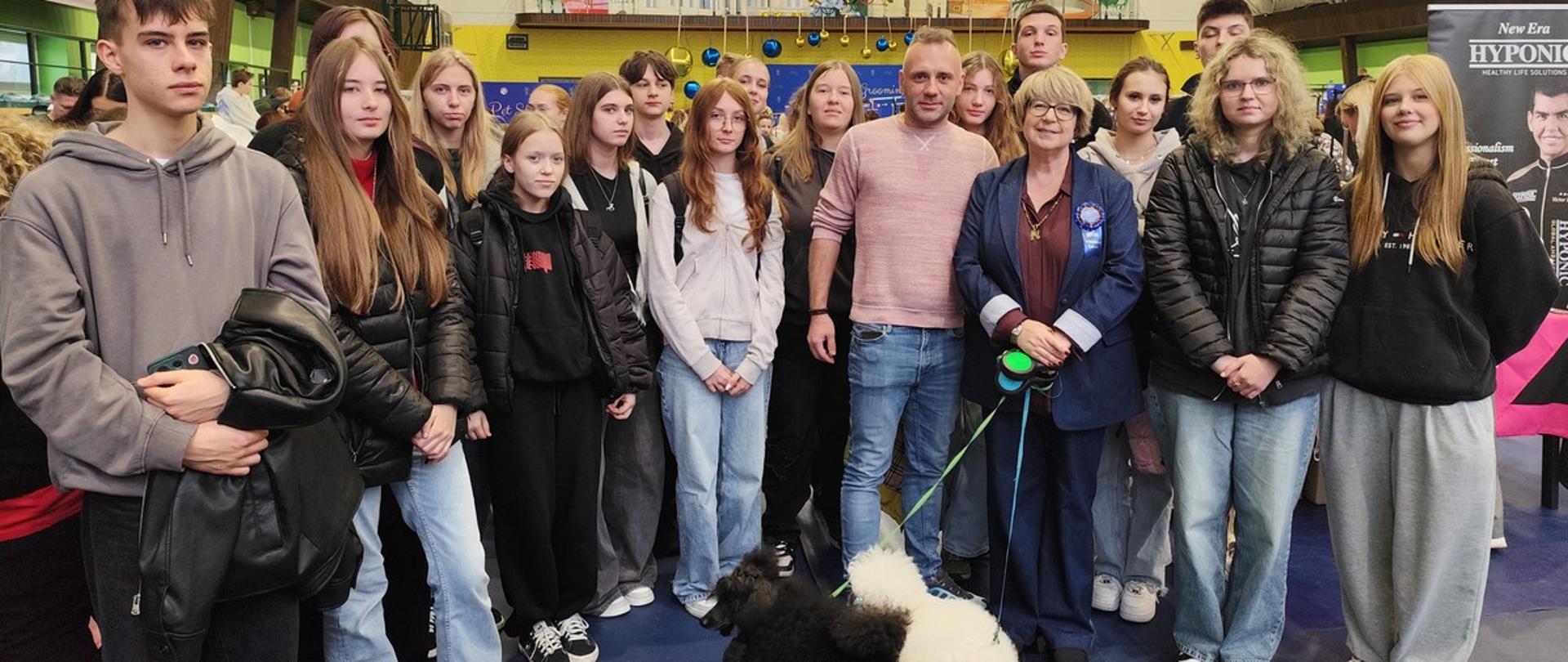 Zdjęcie przedstawia grupę około 15 osób, w różnym wieku stojących w luźnej formacji, pozujących do zdjęcia w dużej, zadaszonej hali. Przed nimi stoją dwa psy - jeden biały drugi czarny.