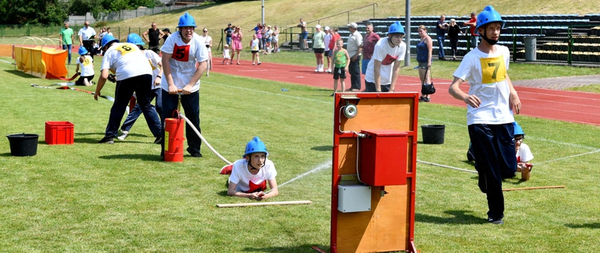 Na pierwszym planie dzieci biorące udział w zawodach napełniają wodą nalewak drugi z zawodników pompuje wodę hydronetką, w tle publiczność na bieżni jak również na trybunach. Wszystko odbywa się na murawie stadionu. 