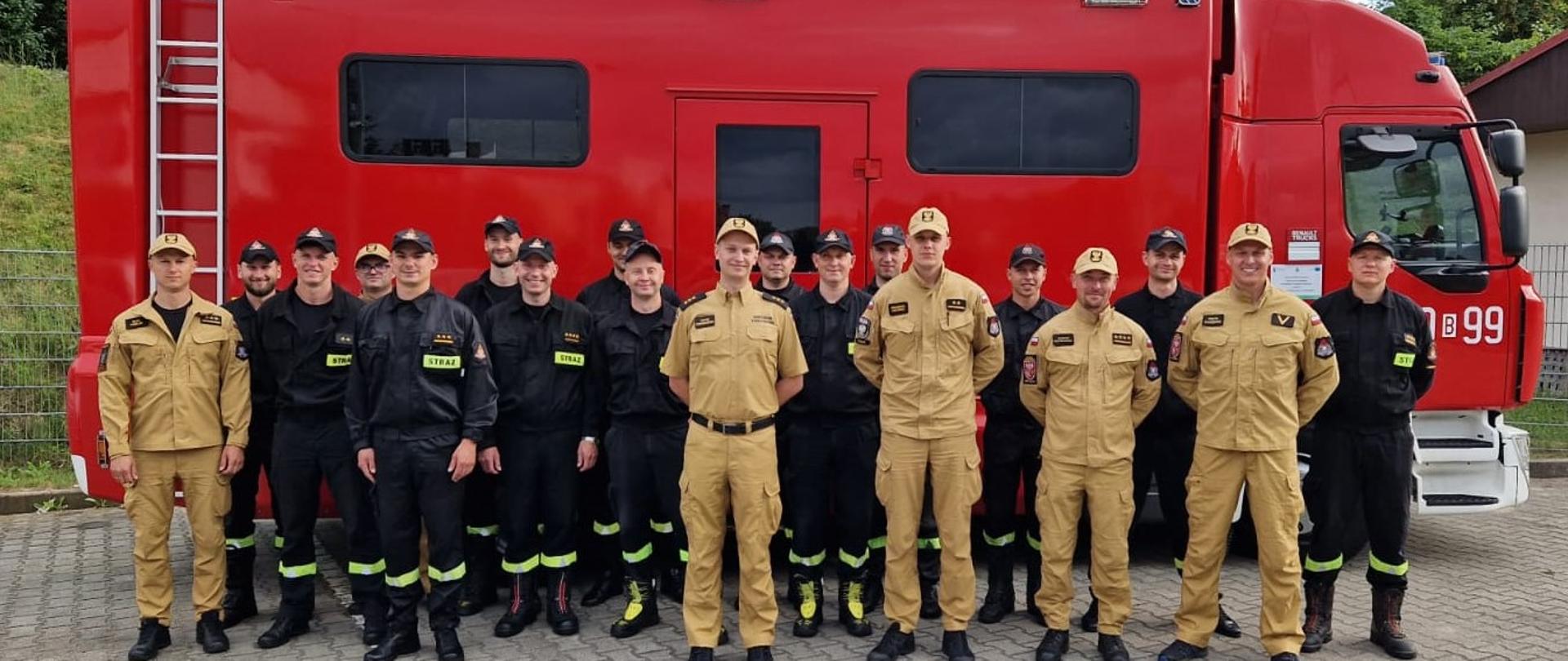 Grupa strażaków w umundurowaniu czarnym i koloru piaskowego stoi na tle samochodu dowodzenia i łączności
