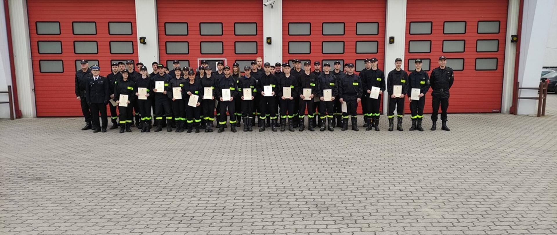 Plac wewnętrzny KP PSP, grupowe zdjęcie kursantów na tle garaży, kursanci prezentują świadectwa ukończenia szkolenia