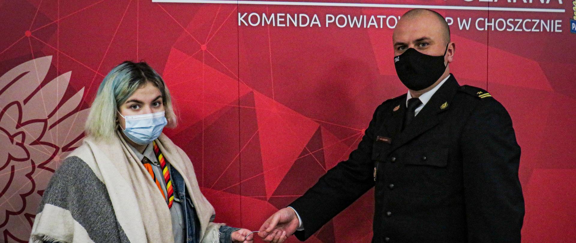 Na fotografii widzimy komendanta powiatowego psp w Choszcznie oraz harcerkę, którzy znajdują się na czerwonym tle z logiem psp. W rękach przekazują sobie lampion ze światłem pokoju.