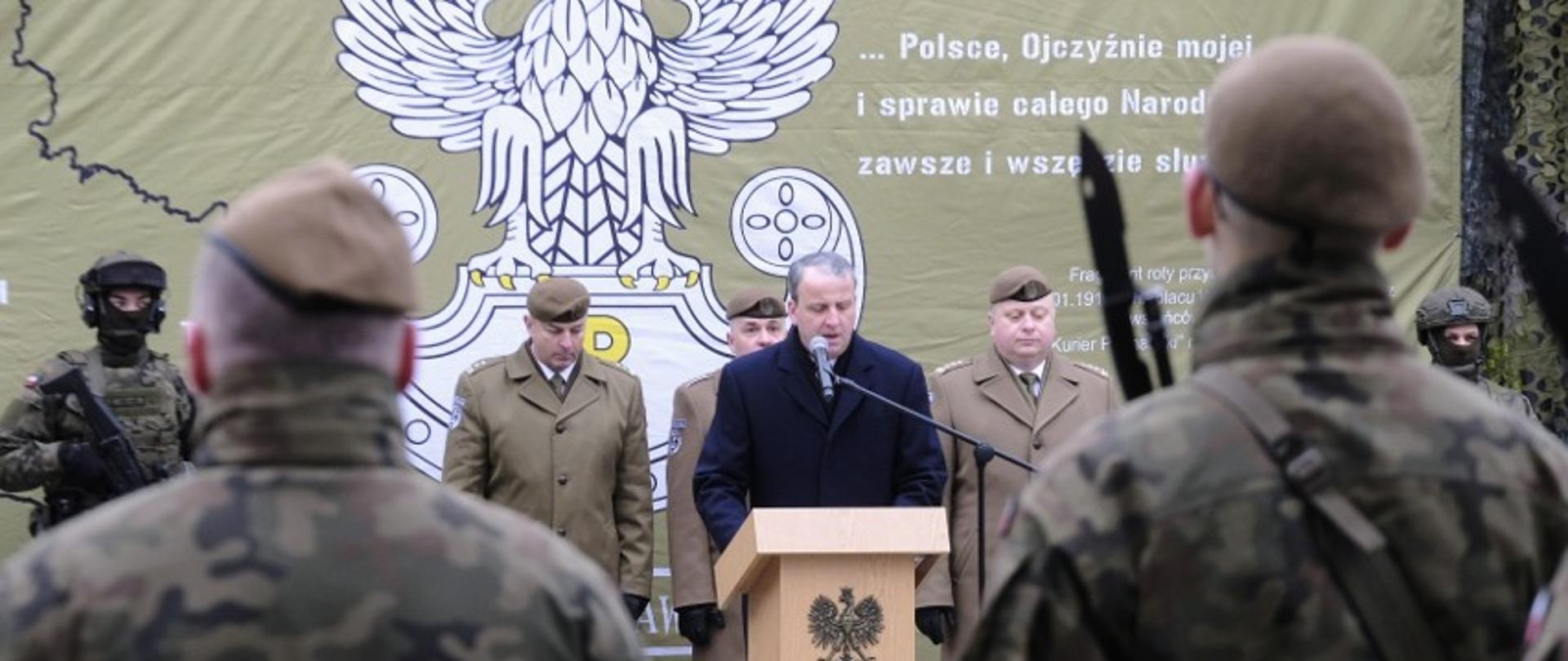 męzczyzna stoi przy mównicy przed grupą żołnierzy, za nim stoją żołnierze w szeregu, w tle na zielonej ściance biały orzeł