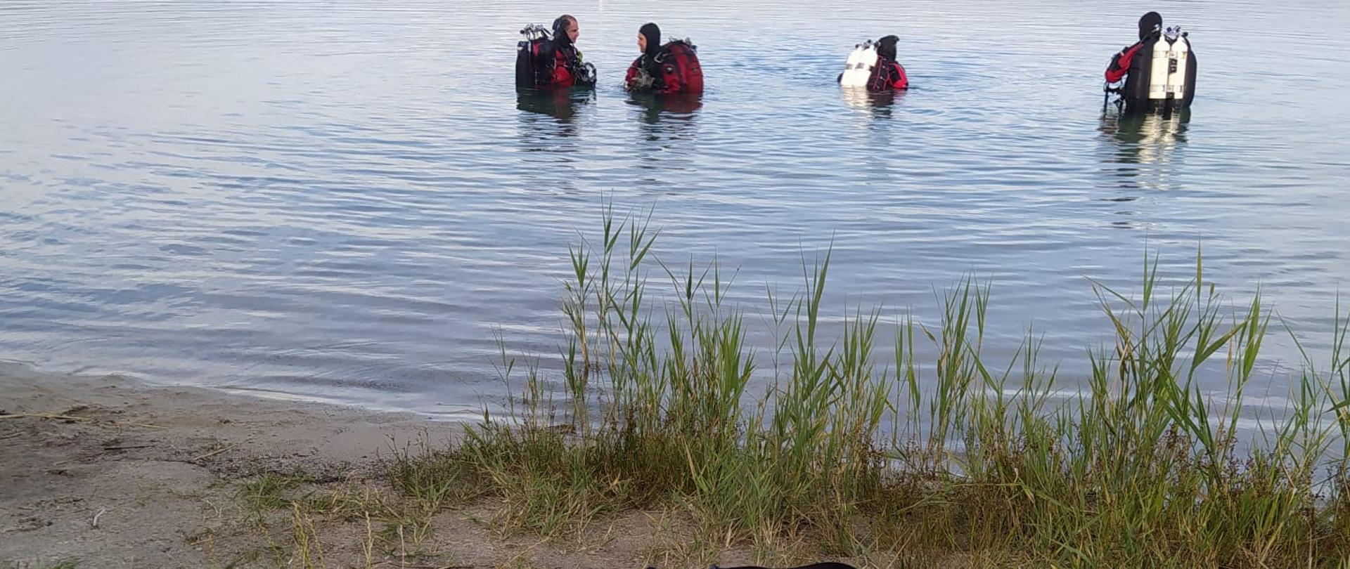Na brzegu torba do udzielania pierwszej pomocy, w tle akwen wodny z widocznymi ratownikami w ubraniach i aparatach.