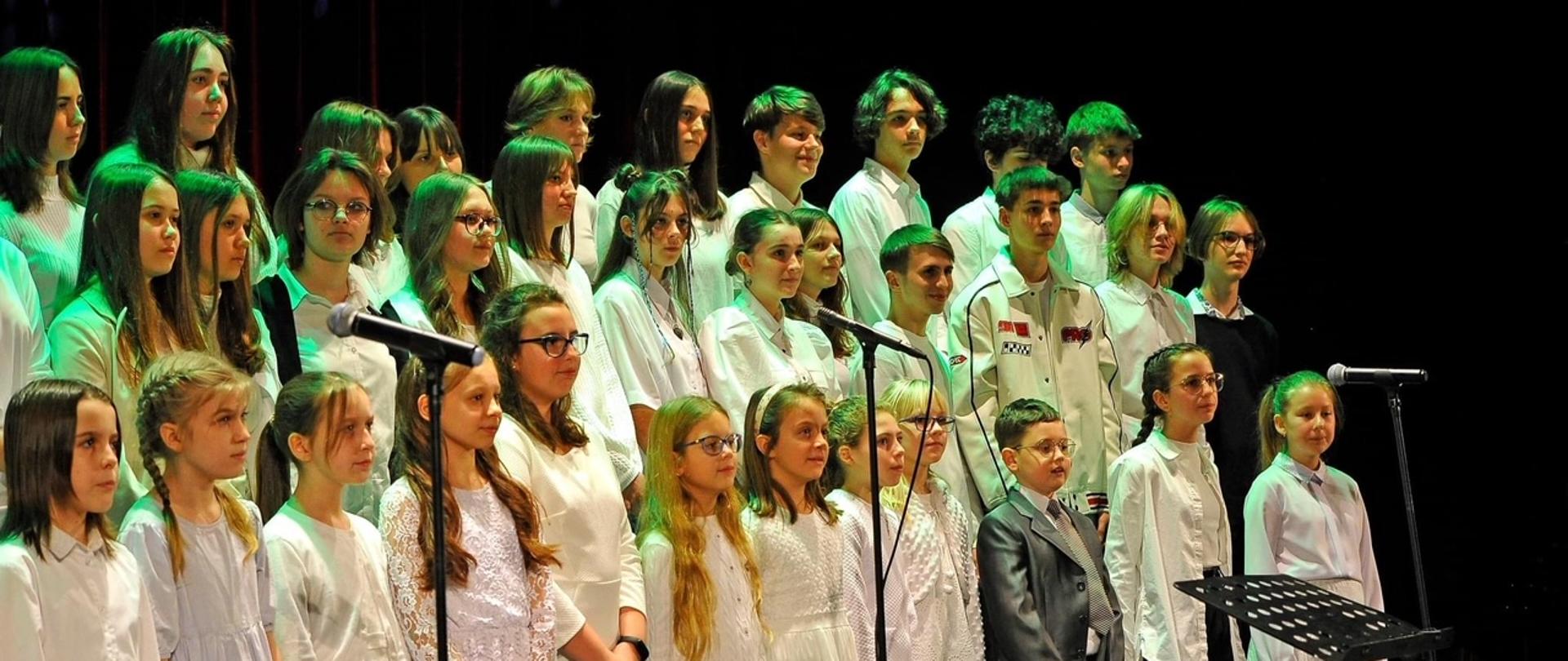 Zdjęcie wykonane na scenie w Kłodzkim Ośrodku Kultury podczas Koncertu Kolęd - 21 stycznia 2023. Na zdjęciu grupowe zdjęcie uczniów ubranych w białe stroje.