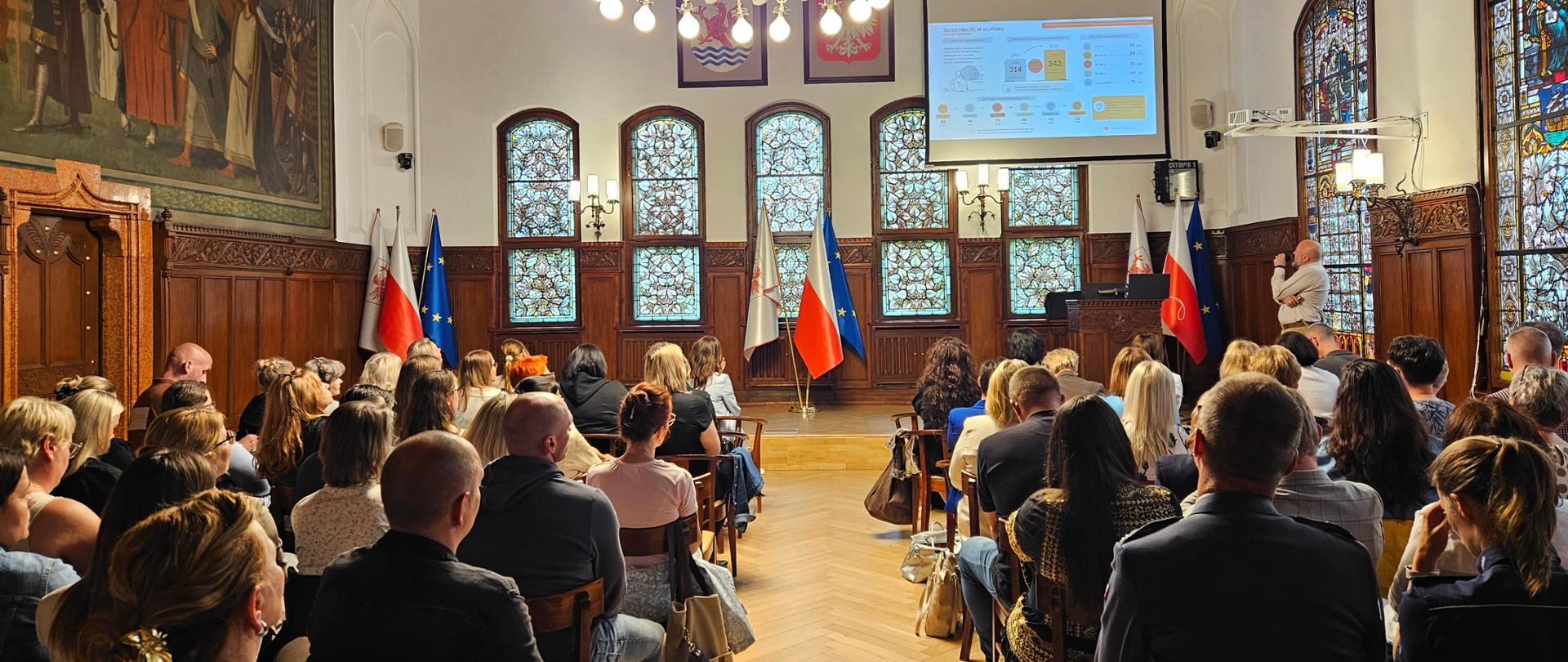 Zdjęcie przedstawia konferencję. W rzędach krzeseł, tyłem do obiektywu, siedzi kilkadziesiąt osób słuchających prezentacji. Na przedniej ścianie wisi ekran projekcyjny wyświetlający slajd z wykresami i danymi.