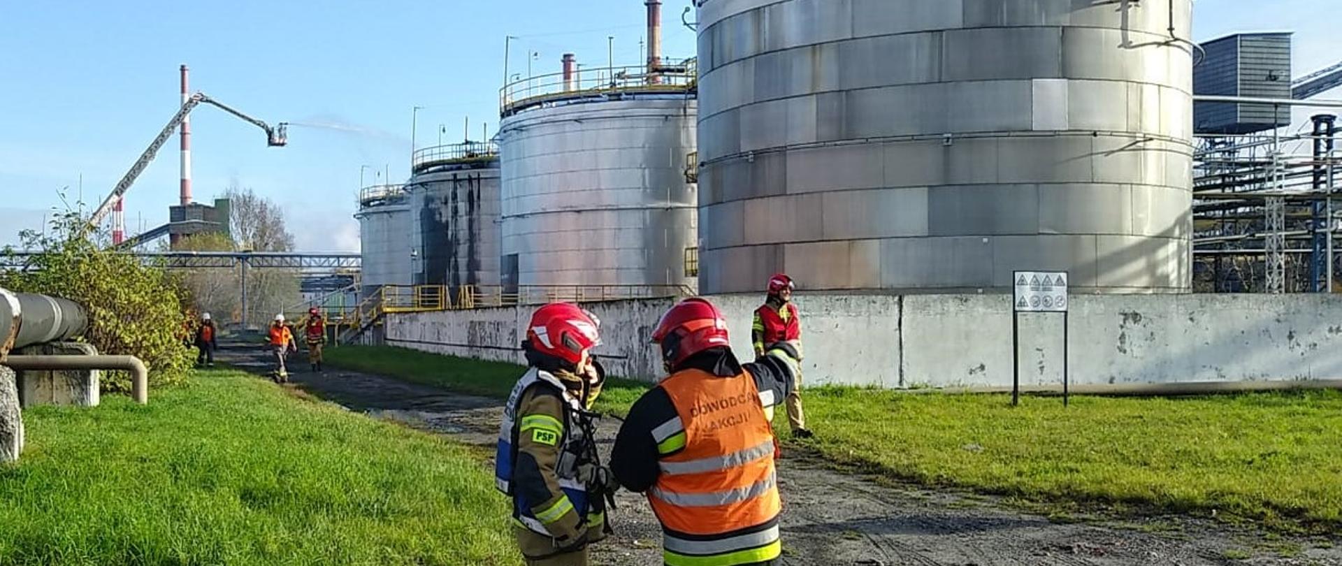 Dowódca jednostki ratowniczej rozmawia z kierującym działaniem ratowniczym Państwowej Straży Pożarnej podczas ćwiczeń na zakładzie dużego ryzyka wystąpienia poważnej awarii przemysłowej. Strażacy w umundurowaniu bojowym. W tle widoczne zbiorniki stokażowe. Na jeden ze zbiorników podawane jest woda z podnośnika ratowniczego.