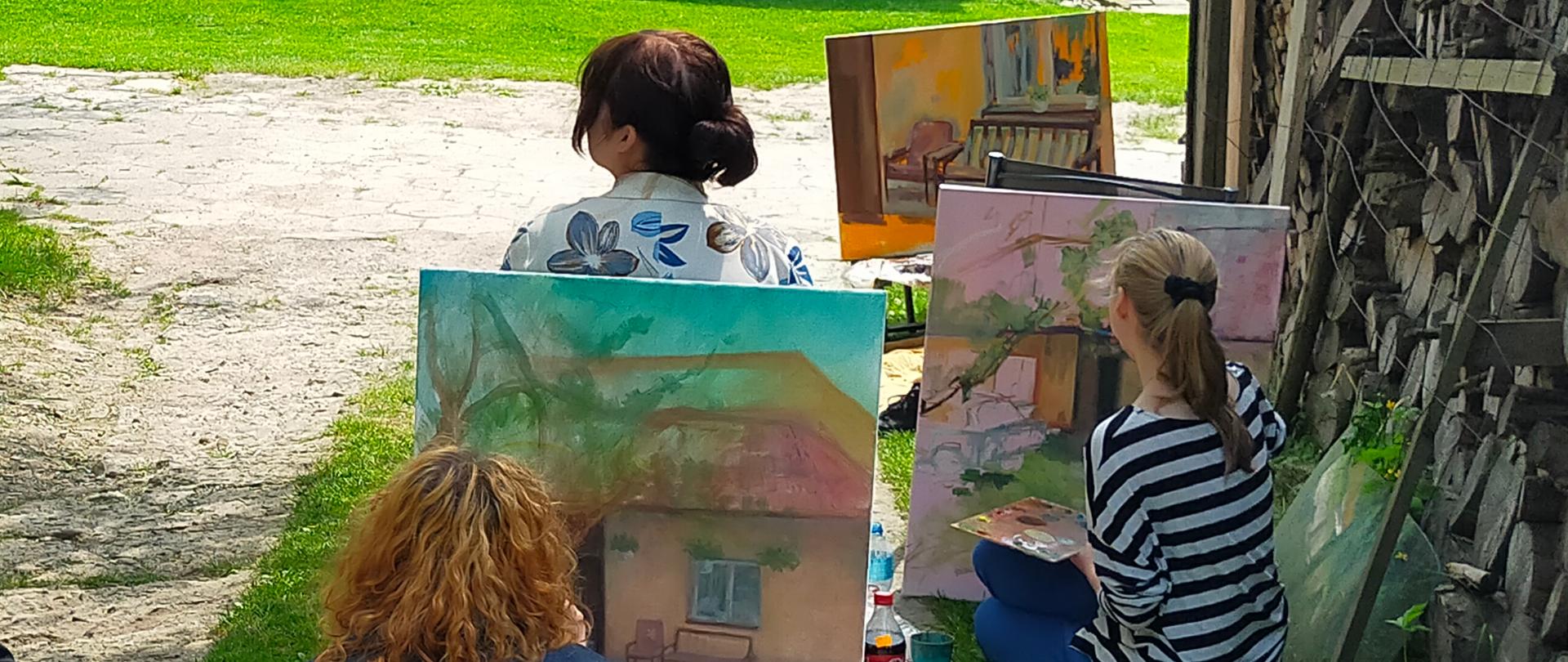 Trzy uczennice siedzą i malują na płotkach, na kocach farby, obok stoi nauczyciel