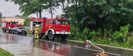 Na mokrej drodze stoją trzy pojazdy strażackie i samochód osobowy. Strażacy rozwijają węże strażackie i używają przenośnej pompy. W tle widać zieleń przydrożnych drzew.