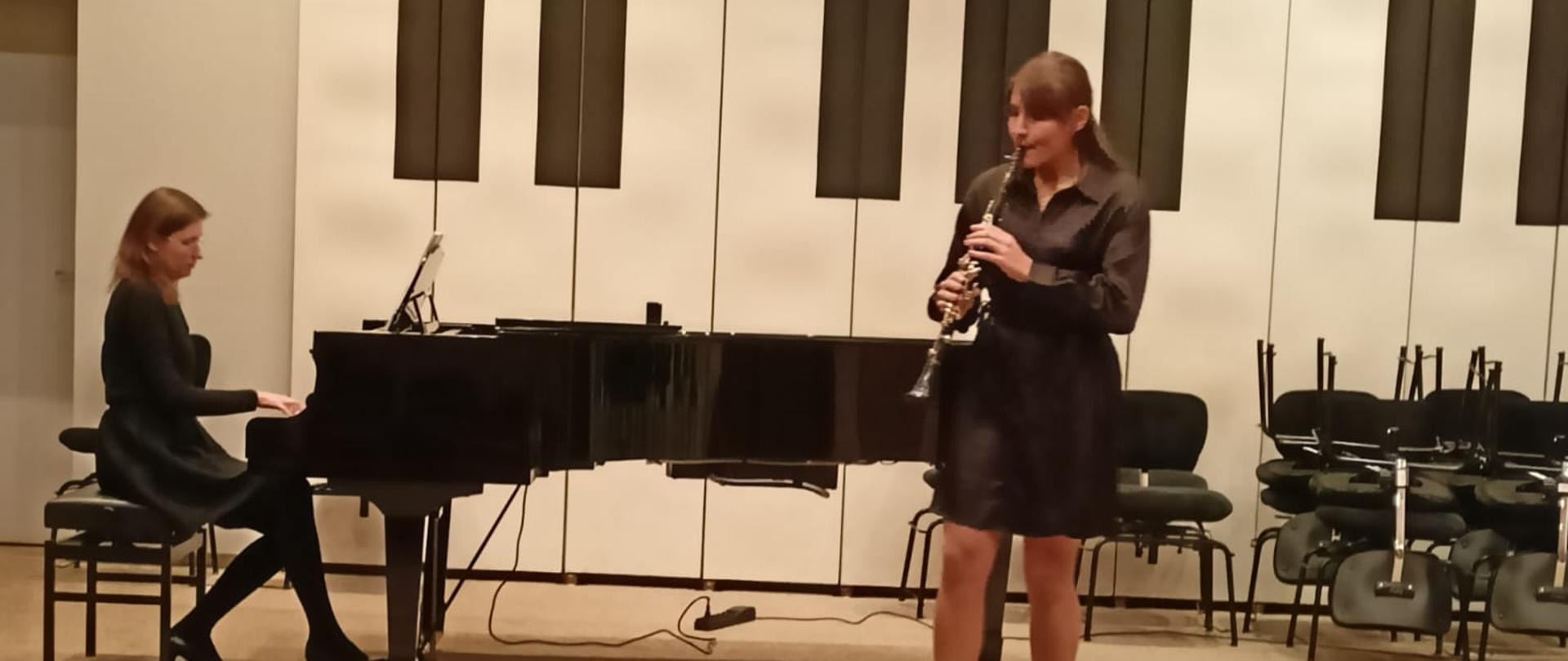 Nastolatka stojąc na estradzie auli PSM gra na klarnecie, za nią na fortepianie gra kobieta, z przodu widać od tyłu fragment widowni z publicznością.
