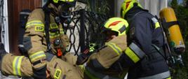 Ćwiczenia w Pakosławiu. Dwóch strażaków wyposażonych w aparaty powietrzne ewakuuje innego strażaka, który uległ wypadkowi wewnątrz budynku. Poszkodowany jest znoszony po schodach zewnętrznych. W tle wejście do budynku.