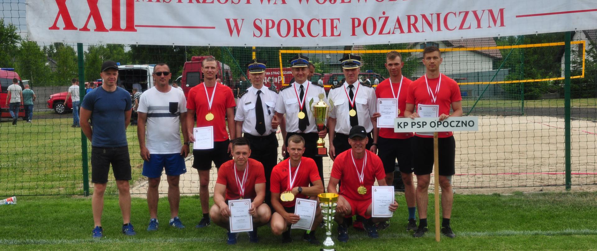 Jedenastu uśmiechniętych strażaków pozuje do zdjęcia stojąc w dwóch rzędach na boisku piłkarskim, ubrani w stroje sportowe z medalami i dwoma pucharami. W środku Komendanci w umundurowaniu służbowym. Nad wszystkimi duży napis w kolorze czerwonym na białym tle o treści dwudzieste drugie mistrzostwa województwa łódzkiego w sporcie pożarniczym. 