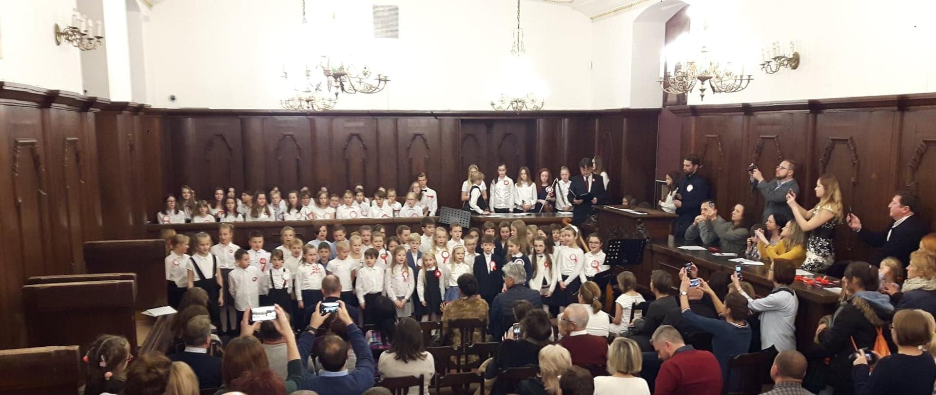 Zdjęcie z koncertu uczniów szkoły muzycznej, przedstawia salę Ratusza Miejskiego z zabytkową dębową boazerią. W centrum zdjęcia chórek dziecięcy w galowych strojach, na pierwszym planie widoczna od tyłu zgromadzona publiczność.