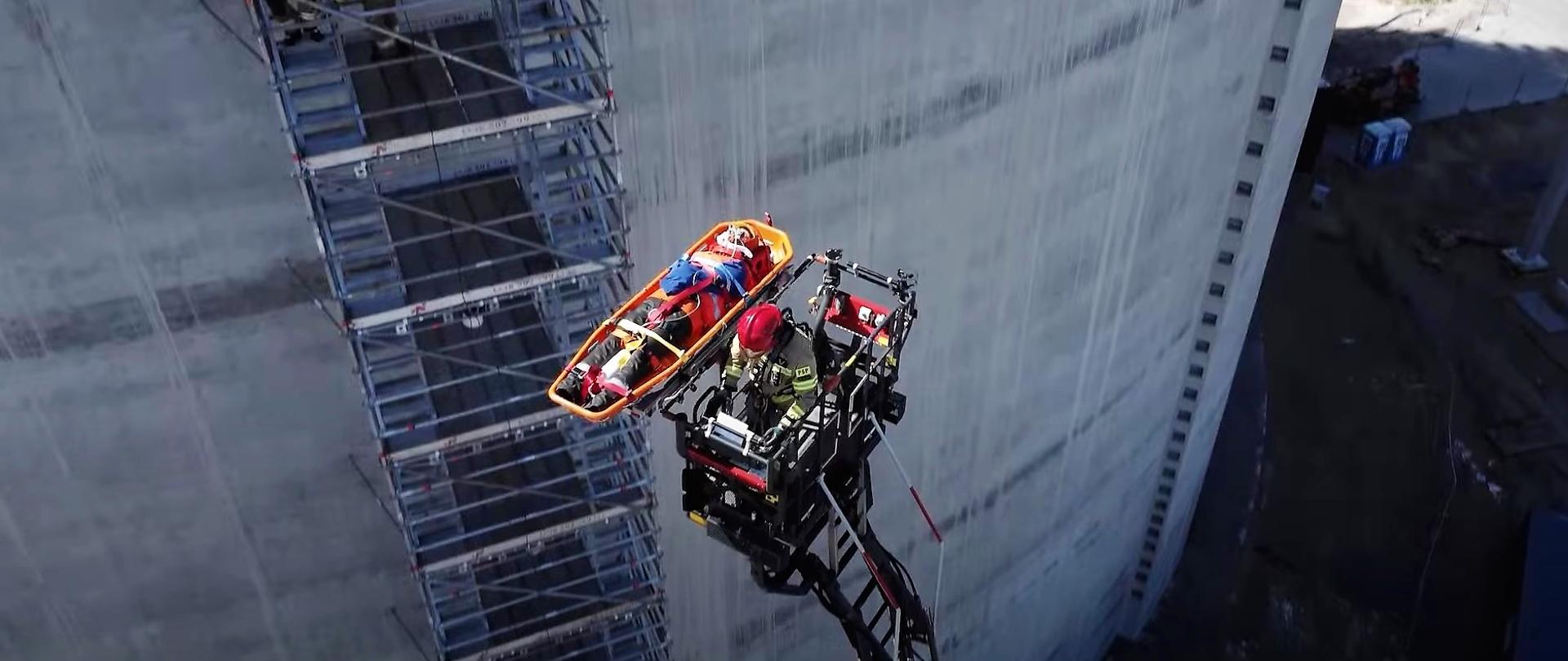 Widok z dużej wysokości na ścianę będącego w budowie zbiornika LNG. Widoczna zewnętrzna ażurowa klatka schodowa przylegająca z zewnątrz do ściany zbiornika a także kosz rozstawionej strażackiej drabiny mechanicznej. Znajduje się w nim strażak w mundurze bojowym. Do konstrukcji kosza przymocowana jest platforma z noszami w których leży osoba poszkodowana ewakuowana ze ściany zbiornika.