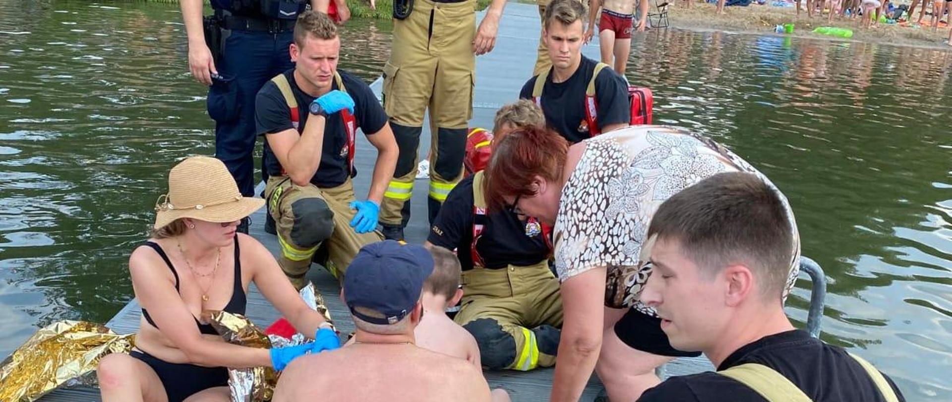 Zdjęcie przedstawia ratowników, którzy udzielili pomocy dziecku na pomoście, nad jeziorem. W tle plaża.