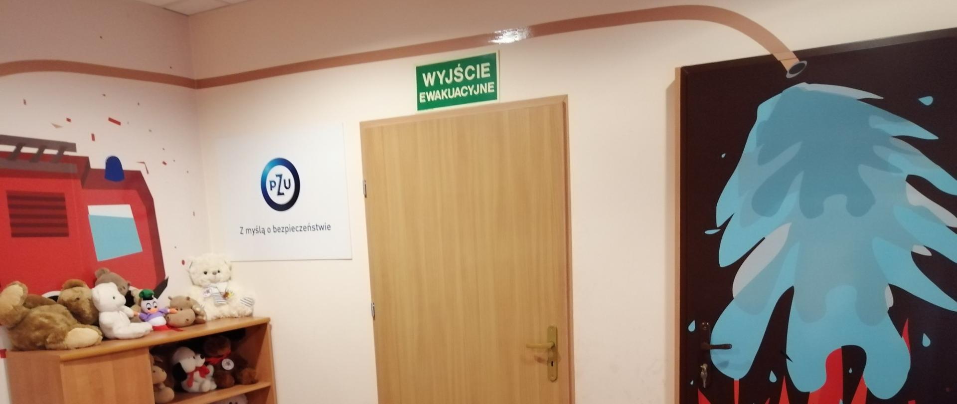 Zdjęcie przedstawia wnętrze sali edukacyjnej Ognik. Widoczne "gorące drzwi" oraz wyjście ewakuacyjne. Na ścianach rysunki o tematyce pożarniczej.
