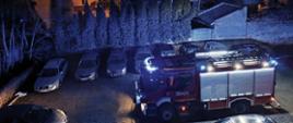 widok z góry samochodu pożarniczego w porze nocnej włączone światła reflektorów, z tyłu drzewa i kamienica mieszkalna oraz samochody osobowe