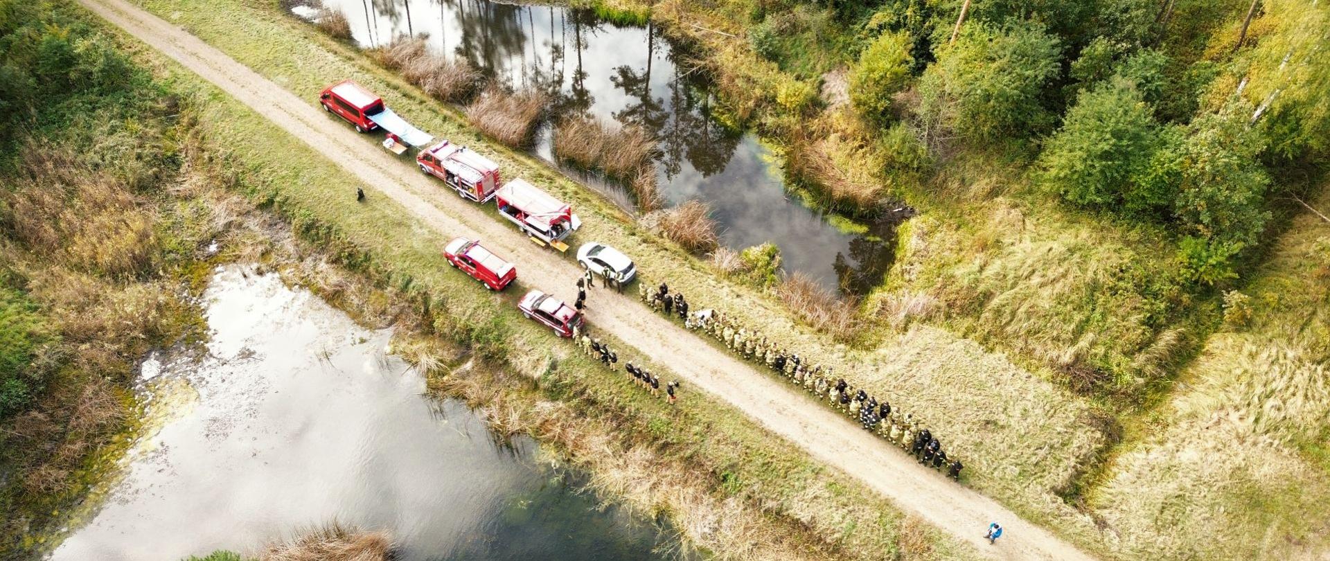 Widok z lotu ptaka przedstawiający wąską, szutrową drogę przebiegającą przez podmokły i zalesiony teren, otoczoną z obu stron wodą. Na drodze, w rzędzie, ustawione są pojazdy ratownicze obok których stoi grupa uczestników ćwiczeń powiatowych.