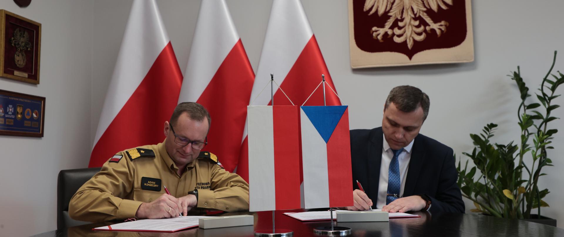 Zastępca komendanta głównego PSP oraz przewodniczący czeskiej części grupy roboczej podpisują dokumenty , na stole flagi Polski i Czech, w tle trzy flagi Polski