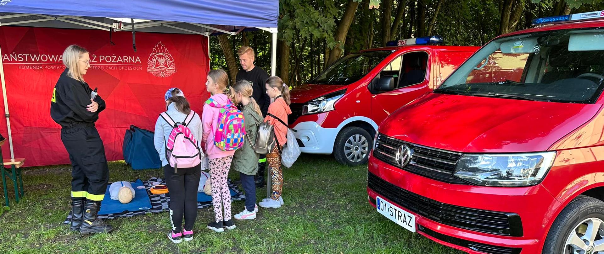 Na zdjęciu znajdują się dzieci stojące przy namiocie pokazowym ze straży pożarnej. Pod namiotem stoi druhna i druh OSP. Obok ustawione są dwa samochody strażackie.