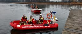 Fotografia przedstawia ujęcie czerwonej łodzi ratowniczej na wodzie w której siedzi trzech strażaków w kaskach, kamizelkach asekuracyjnych i w ubraniach specjalnych. W prawym dolnym rogu kadru widoczny pomost. W tle widoczna inna łódź ratownicza, dalej widoczny brzeg jeziora. Jest dzień.