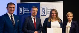 Na tle baneru z napisami Polska Komisja Akredytacyjna stoją od lewej mężczyzna w granatowym garniturze, minister Wieczorek, trzymająca otwarty dokument kobieta w czarnej marynarce i wiceminister Mrówczyńska.