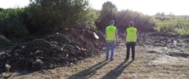 Dwóch inspektorów w zielonych kamizelkach z napisem „Inspekcja Ochrony Środowiska” w tle hałda nielegalnych odpadów.