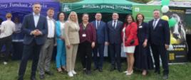 Przed trzema namiotami plenerowymi na trawiastym polu stoi rząd dziesięciu elegancko ubranych osób – kobiet i mężczyzn w garniturach, marynarkach i sukienkach. Po lewej stronie stoi namiot z napisem „NFZ Fundusz zdrowia”, w centrum zielony namiot z hasłem „Rolniku, pracuj bezpiecznie”, a po prawej stronie baner reklamowy. Wszyscy uczestnicy pozują przodem, uśmiechając się do zdjęcia.