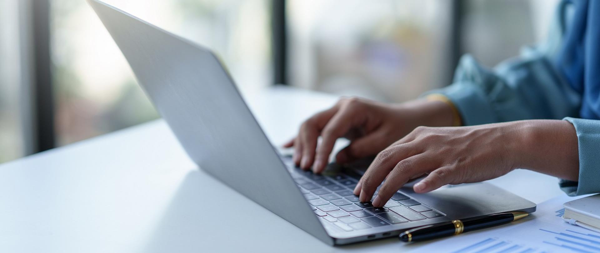 Dłonie na klawiaturze laptopa, który leży na biurku. Obok długopis. 