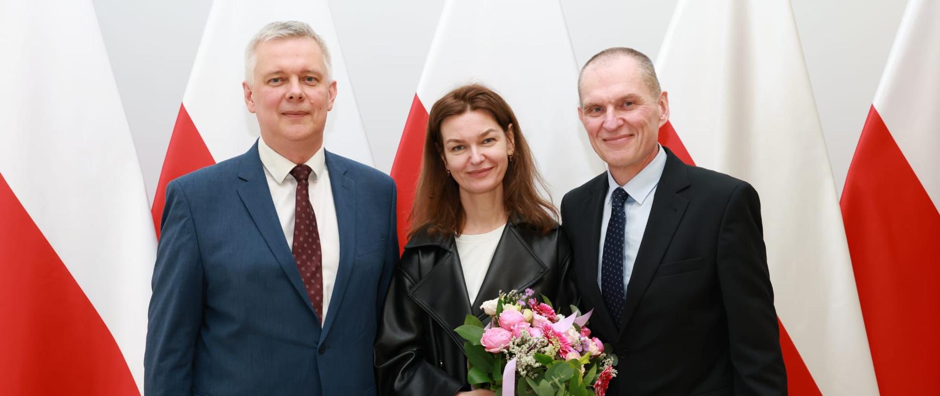 Na zdjęciu trzy osoby: minister Tomasz Siemoniak (po lewej), Oksana Poczobut (w środku) oraz Andrzej Poczobut (po prawej). Oksana Poczobut trzyma w rękach bukiet kwiatów. Osoby stoją na tle 5 flag Polski (biało-czerwonych).