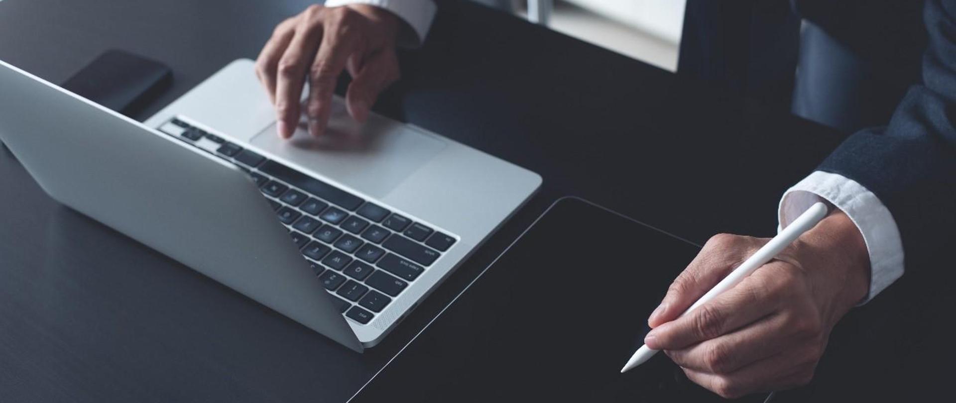 Mężczyzna w garniturze (ujęcie rąk i nóg) stoi przy biurku, jedną rękę trzyma na laptopie, w drugie trzyma rysik nad tabletem