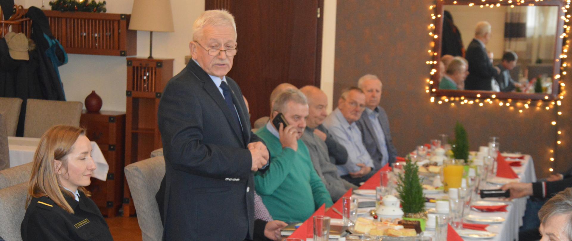 Świąteczne spotkanie strażackich emerytów. Sala w jednej z rawickich restauracji. Po obu stronach stołu zasiadają uczestnicy spotkania. Po lewej stronie siedzi kobieta w mundurze strażackim, a obok niej stoi mężczyzna w ubiorze cywilnym, który przemawia. tuz za nim inny mężczyzna w zielonym swetrze rozmawia przez telefon. Na stole są białe filiżanki, szklanki i dzbanki oraz czerwone serwetki.