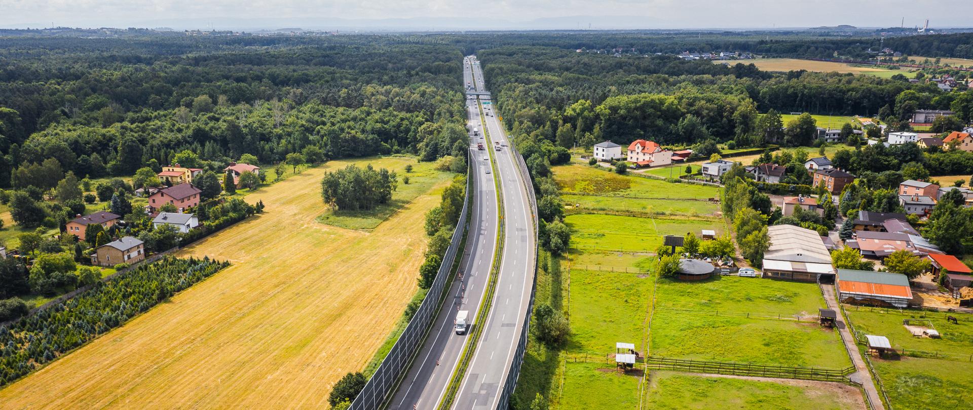 Widok z lotu ptaka na drogę ekspresową S1 przebiegającą między terenami rolnymi, zabudową jednorodzinną i lasami. Wzdłuż trasy znajdują się wysokie ekrany akustyczne, a po jezdni poruszają się samochody ciężarowe i osobowe.
